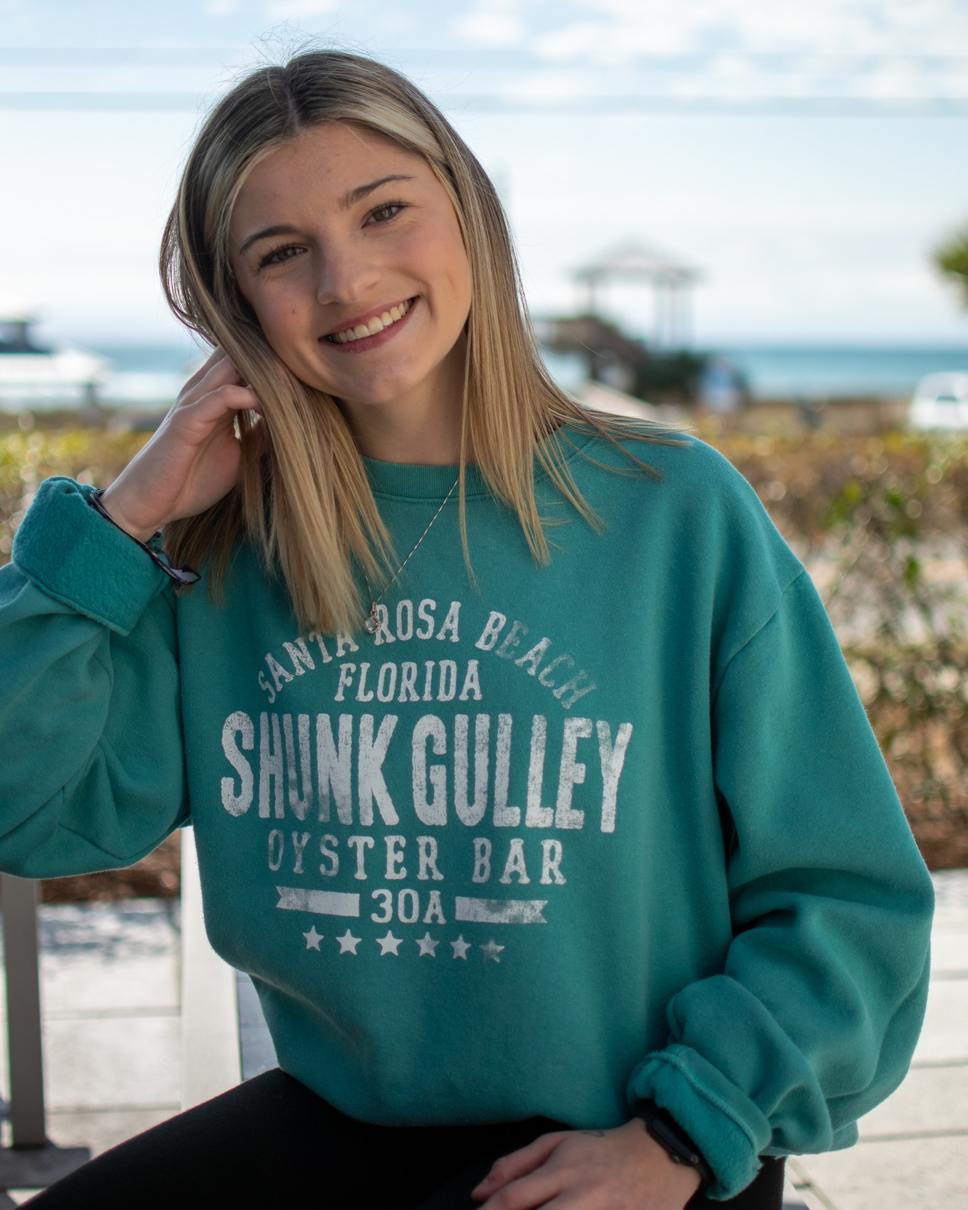 Smiling person in a teal sweatshirt leaning outdoors near the beach with palm trees in the background