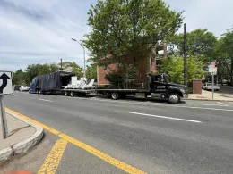 Black flatbed truck hauling equipment on a city street by a brick building and trees