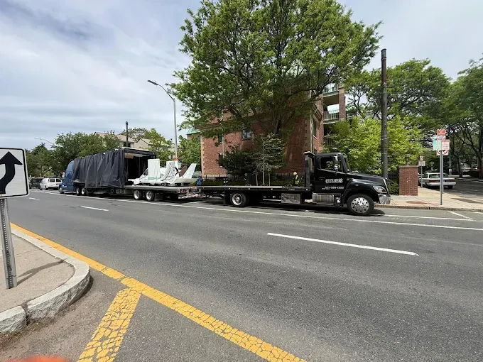 Black pickup truck hauling a flatbed trailer on a city street beside a brick building and trees.