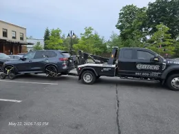 Two damaged vehicles in a parking lot, a black SUV hitched to a tow truck, with trees and a building nearby.