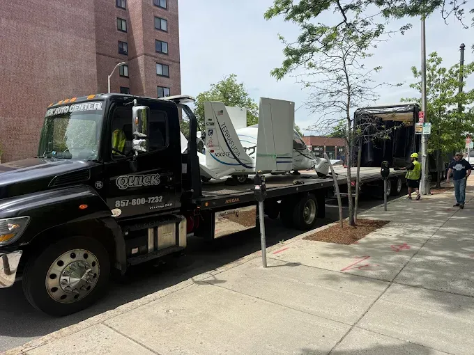 Black flatbed tow truck carrying equipment parked along a city sidewalk with a building and trees nearby