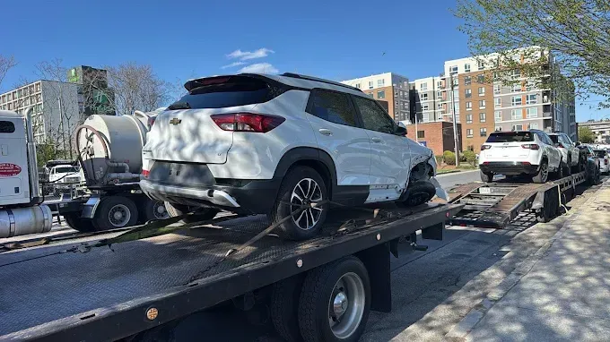 White SUV on a flatbed tow truck in a parking lot, with apartment buildings and a blue sky in the background