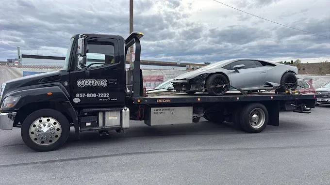 Black tow truck hauling a silver sports car on a flatbed beside a highway.