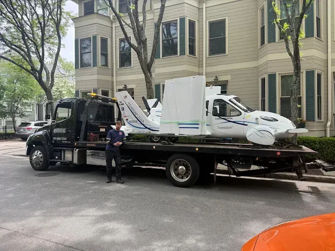 Tow truck hauling a white car on a flatbed in front of a building, with a worker beside it.