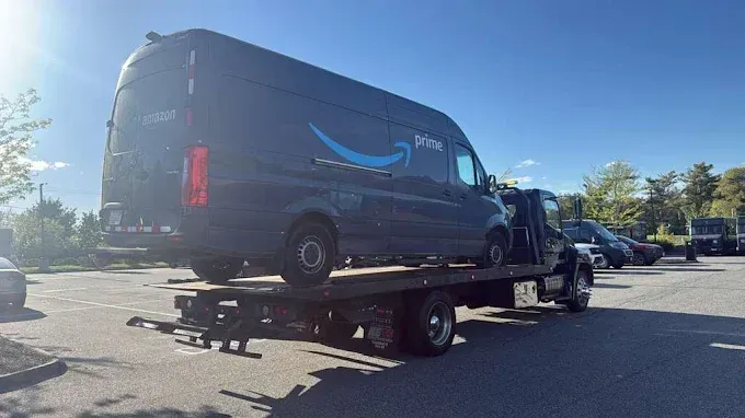 Dark gray Amazon delivery van on a flatbed tow truck in a sunny parking lot