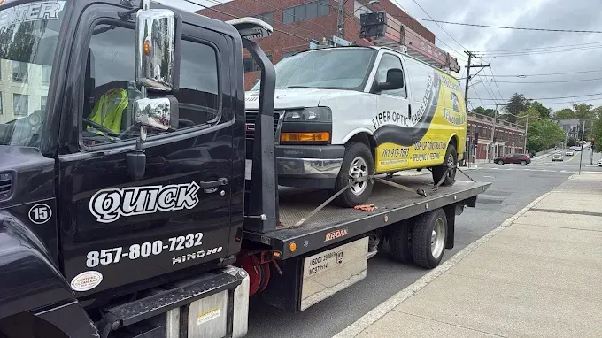 Tow truck carrying a white box truck on a city street beside a sidewalk and brick buildings