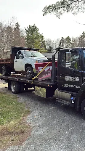 Tow truck hauling a white SUV on a flatbed beside a wooded gravel road