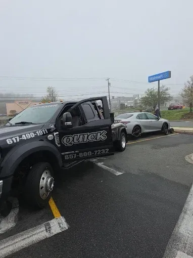 Black tow truck hauling a silver sedan in a wet parking lot under gray skies