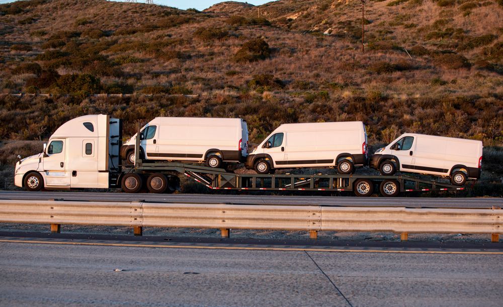 Four white semi trucks parked in a line beside a highway, with a hillside in the background