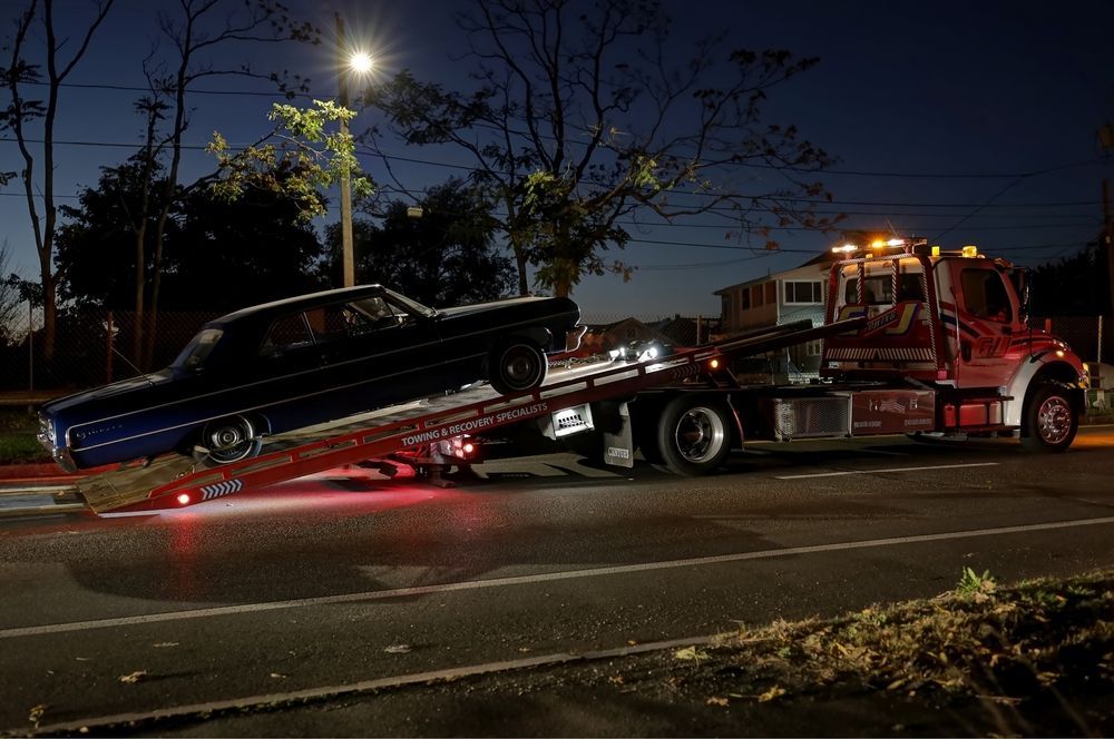 Tow truck hauling a black car on a trailer at night on a city street under streetlights