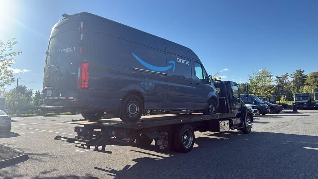 Dark Amazon delivery van on a flatbed tow truck in a parking lot under a clear blue sky