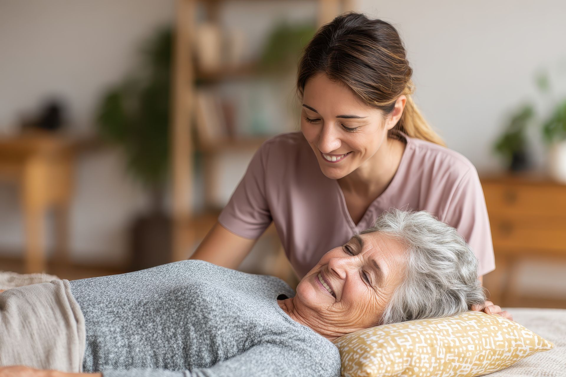 Smiling caregiver attends to senior woman resting on a pillow indoors.