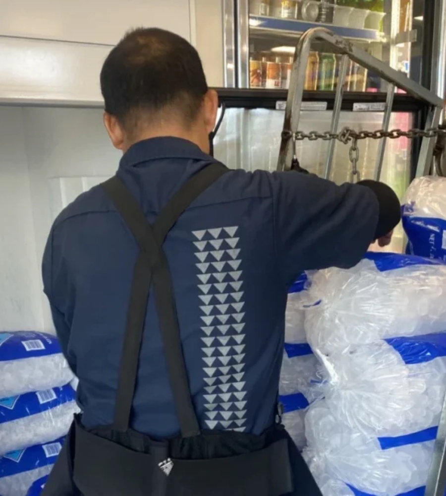 A person wearing a dark blue shirt with triangle patterns on the back stacks bags of ice inside a freezer unit.