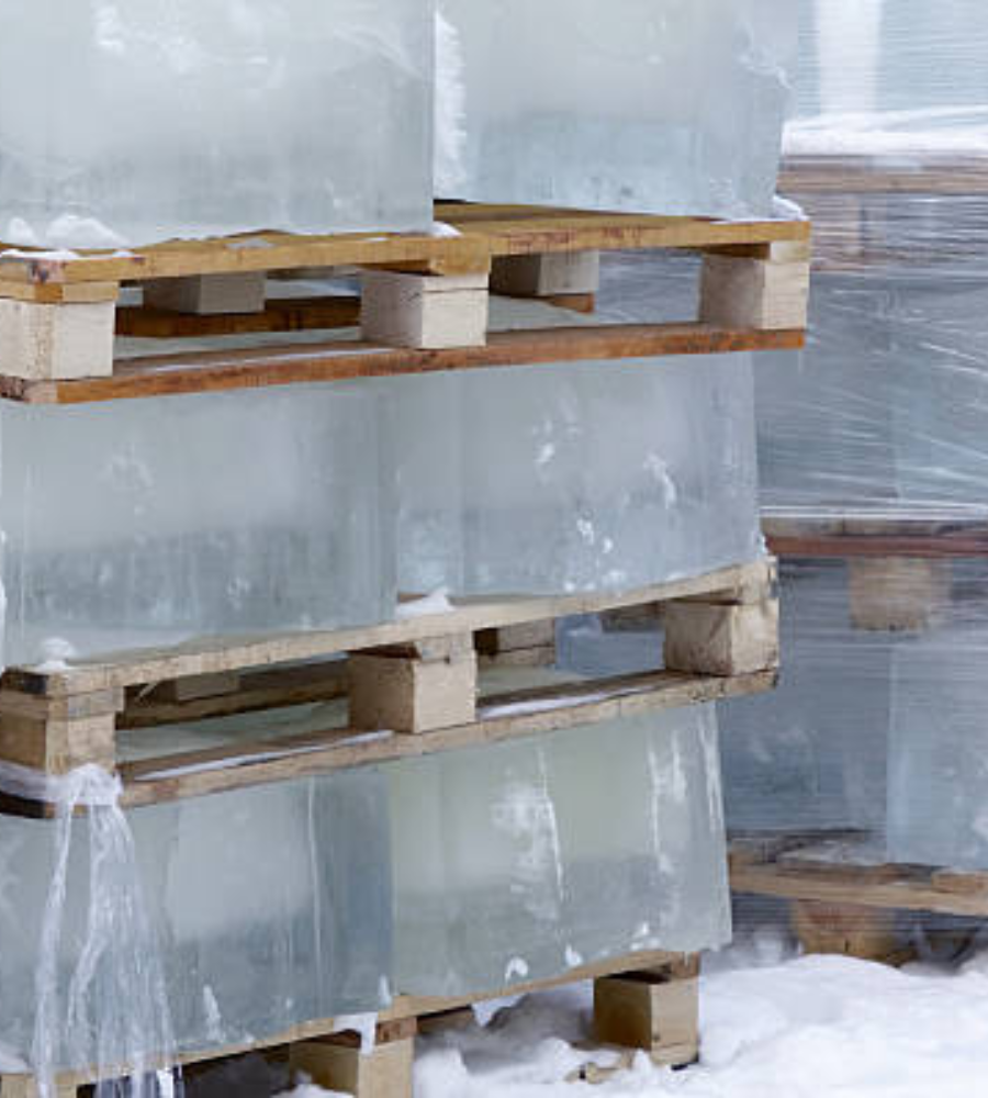 Large, clear blocks of ice stacked on wooden shipping pallets sitting on a snowy ground.