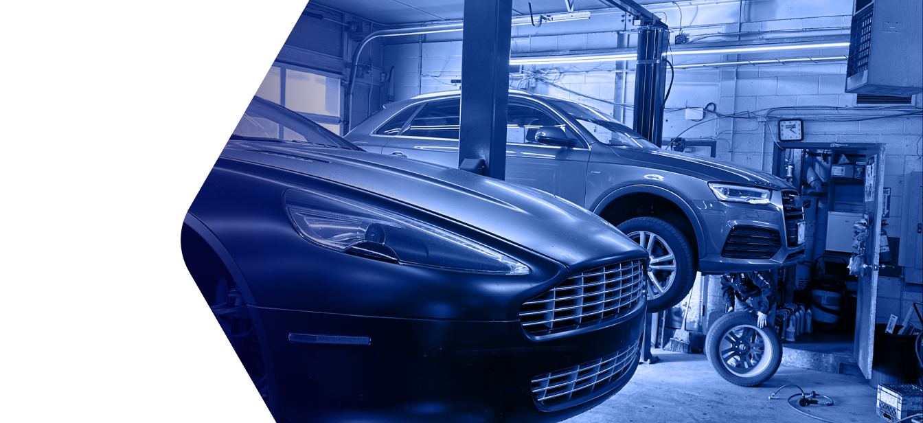 A workshop with an Aston Martin in the foreground and a car on a hydraulic lift in the background, in blue-tinted light. | Heritage Auto & Tire Service