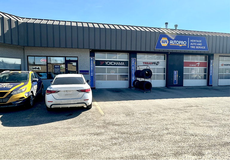 A commercial auto shop exterior with two cars parked in front of three service bay doors under a sunny sky. | Heritage Auto & Tire Service