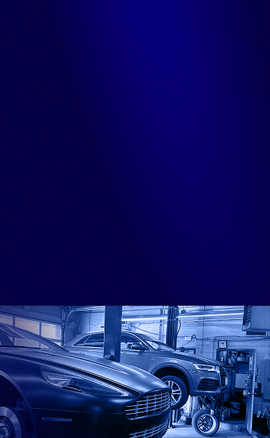 A workshop with an Aston Martin in the foreground and a car on a hydraulic lift in the background, in blue-tinted light. | Heritage Auto & Tire Service