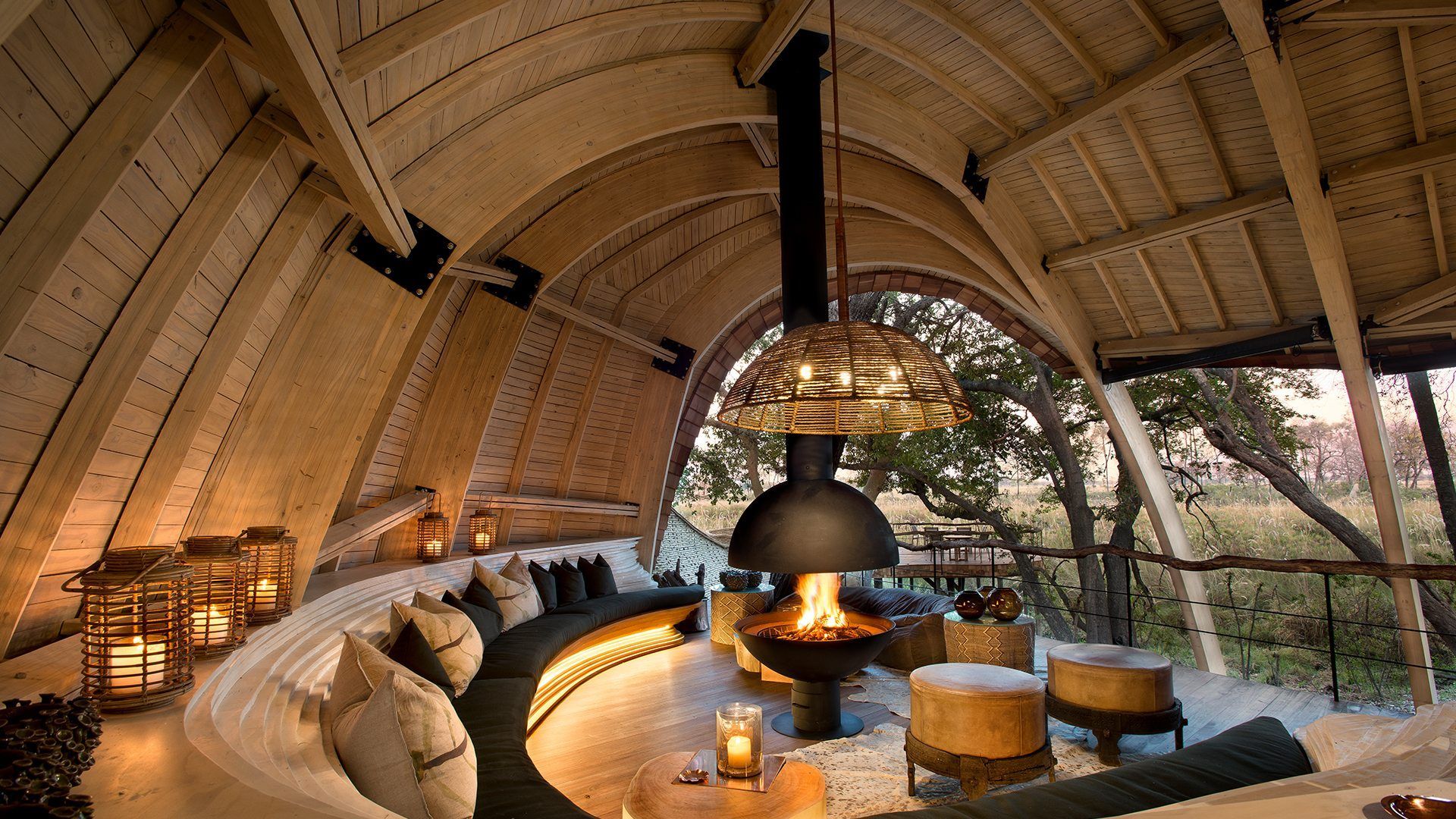 A curved, modern indoor lounge with wooden architecture, a fireplace, and views of a forest landscape.
