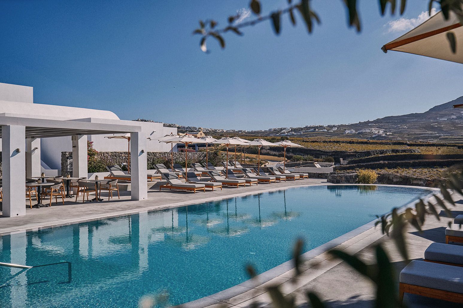 Swimming pool and lounge chairs at a resort with a bright blue sky and distant hills. Vedema, a Luxury Collection Resort, Megalochori
