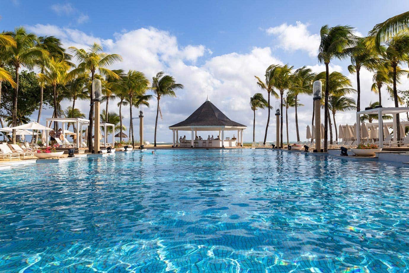 A turquoise swimming pool at a tropical resort, featuring a central gazebo bar, lounge chairs, and tall palm trees. Heritage Le Telfair Golf & Wellness Resort