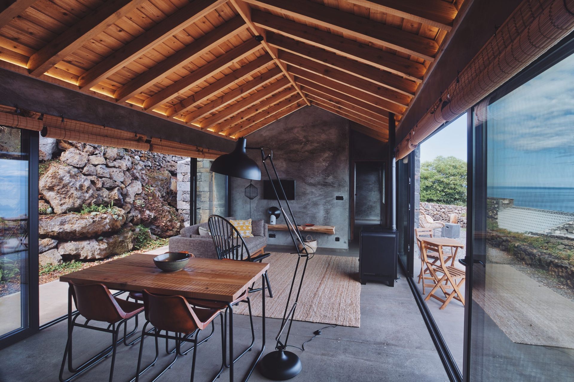 A modern, open-plan cabin interior with a wooden ceiling, stone walls, dining table, and glass walls overlooking the sea.