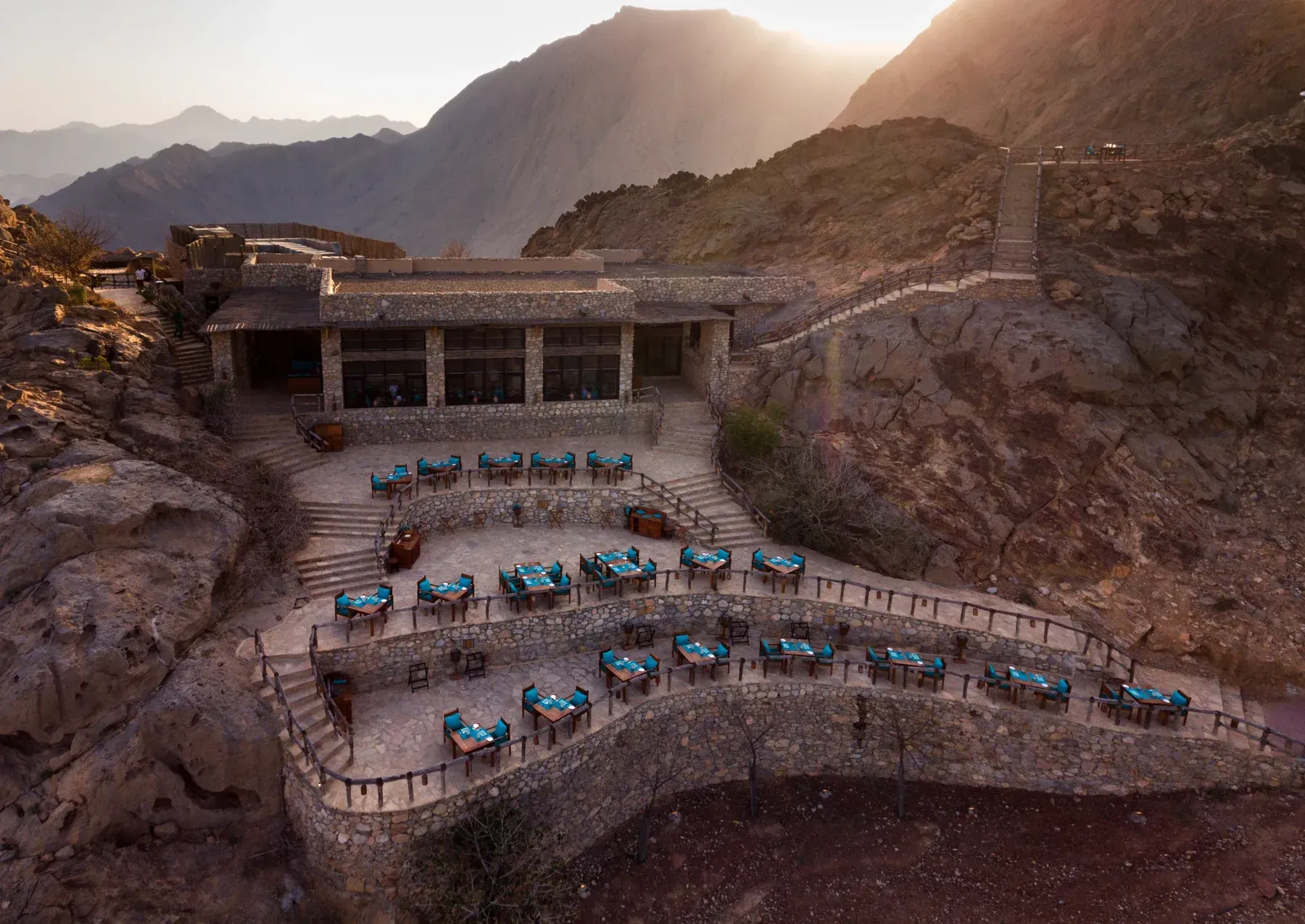 Terraced hillside resort with blue chairs and tables overlooking a rocky mountain valley at sunset Six Senses Zighy Bay Oman
