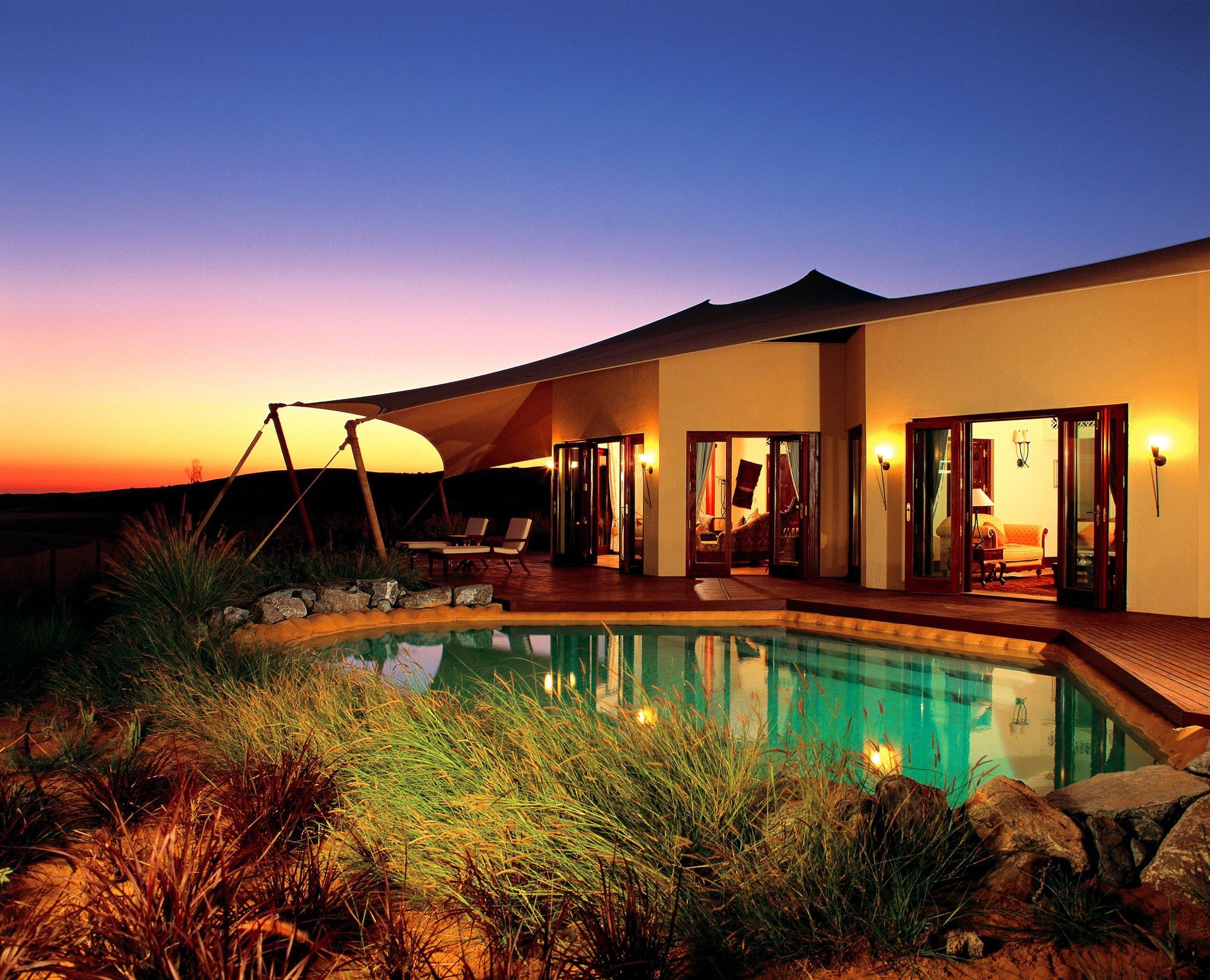 Luxury villa with a pool at sunset. Exterior view with open doors, wooden deck, and a canopy. Al Maha