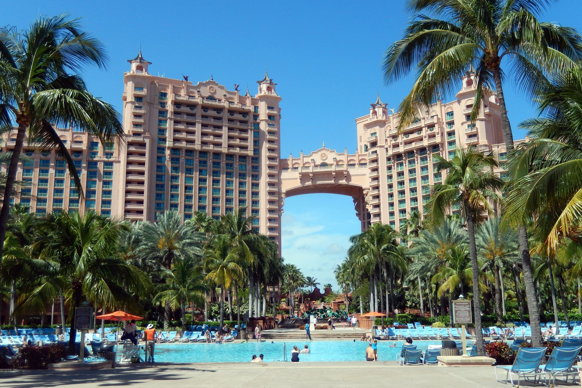 Pink hotel towers connected by a bridge over a pool with palm trees, blue sky. The Atlantis Hotel