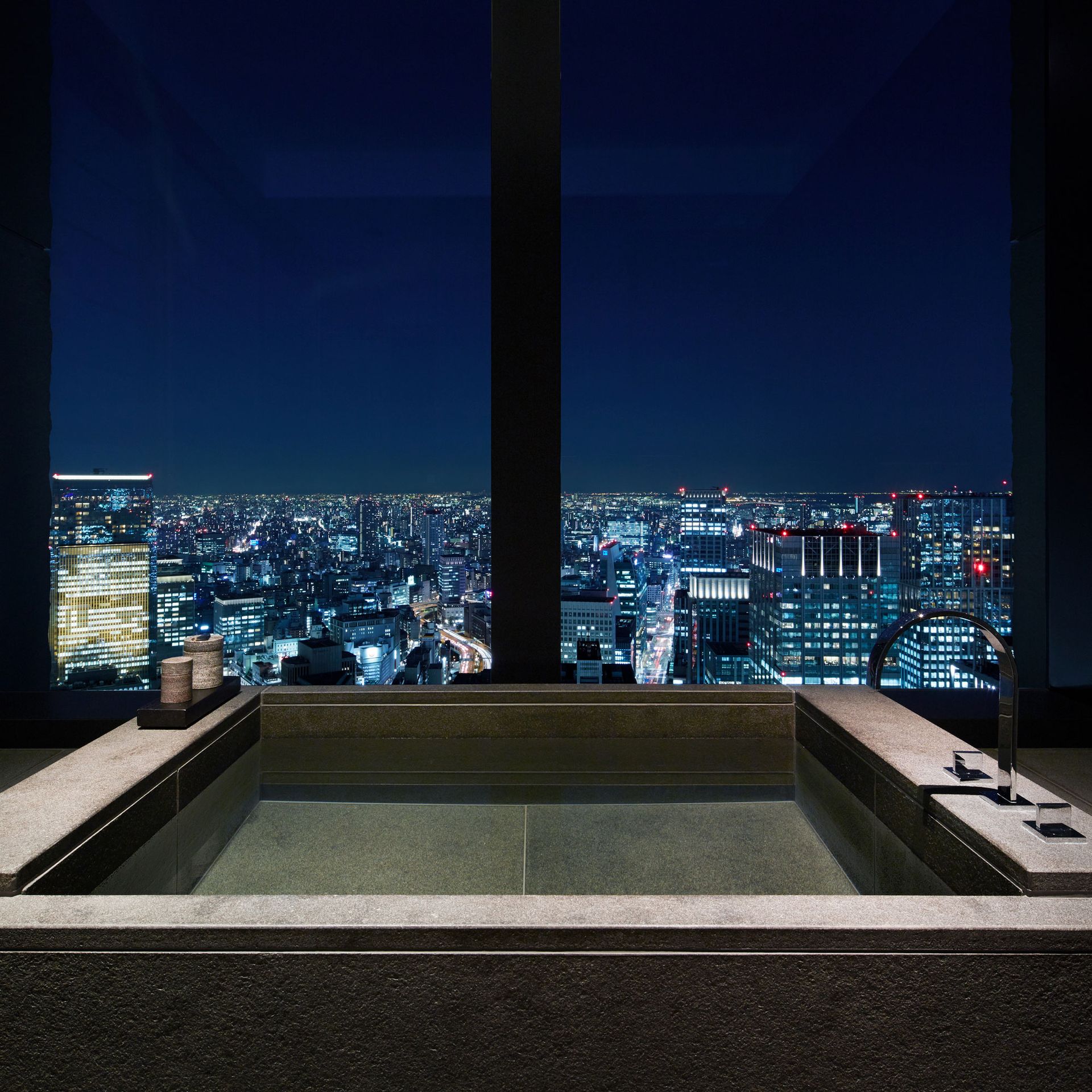 A luxurious bathroom with a cityscape view at night. Features a dark stone bathtub and large window. The Aman Tokyo