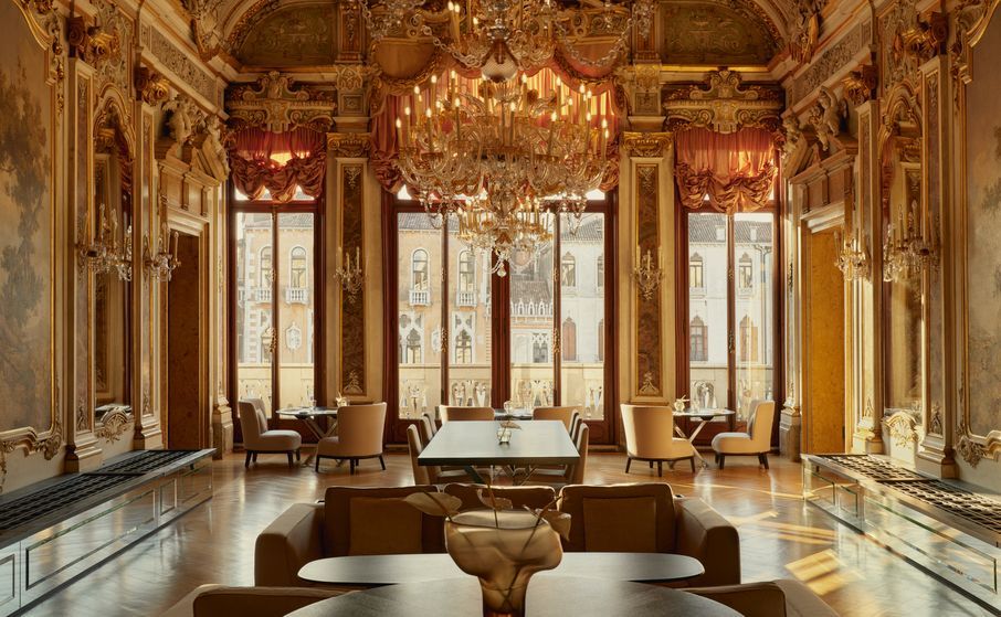 Ornate dining room with golden décor, large windows, chandelier, and modern furniture. Aman-venice_italy_