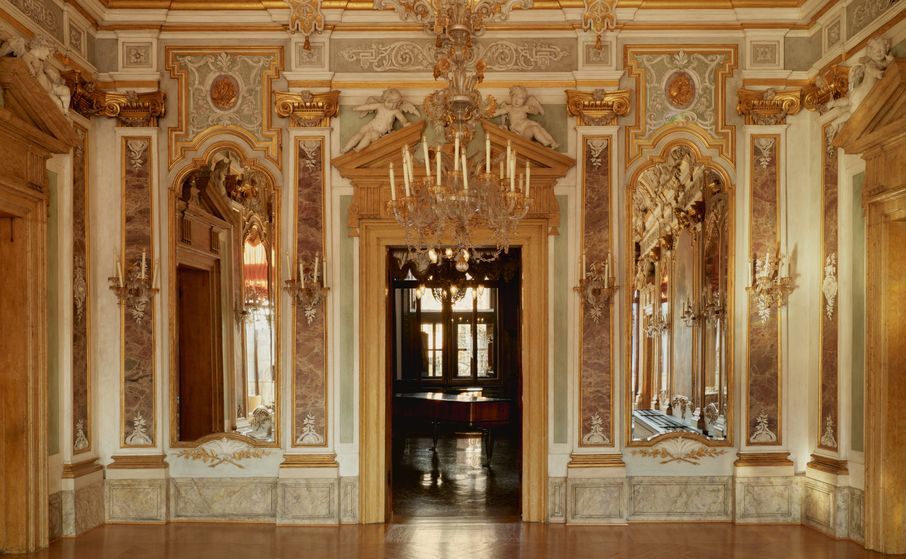 Ornate, gilded room with marble and mirrors; a large chandelier hangs above a doorway. Aman-venice_italy_
