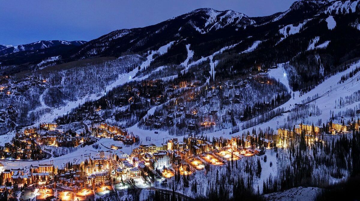 Snowy mountain at dusk, with lights from a town nestled in the valley. Aspen