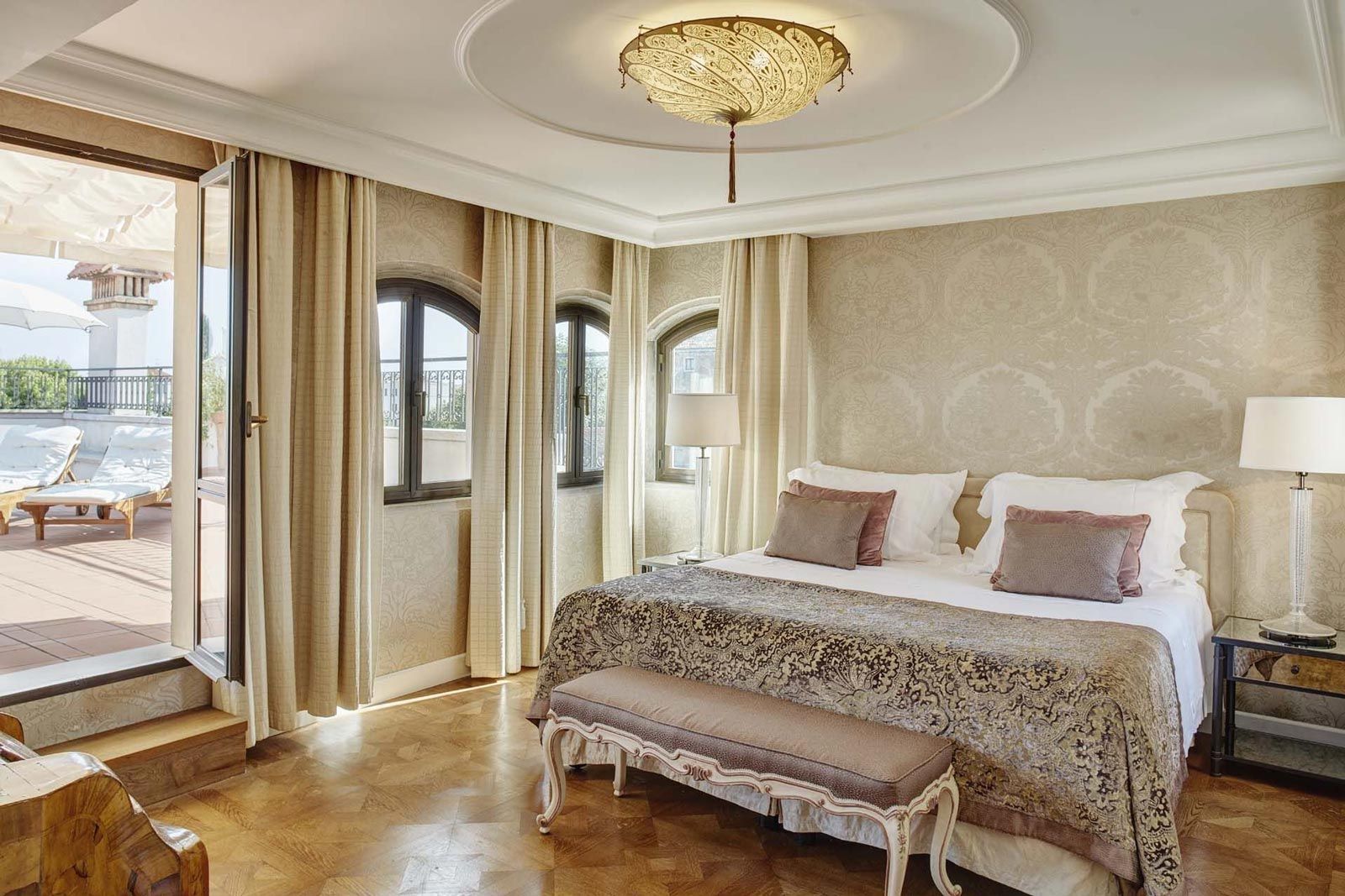Luxury hotel bedroom with bed, terrace access, and ornate ceiling light.