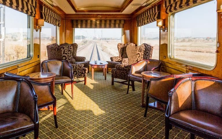 A luxury train observation car with leather chairs and patterned carpets, looking out over tracks through a large window.