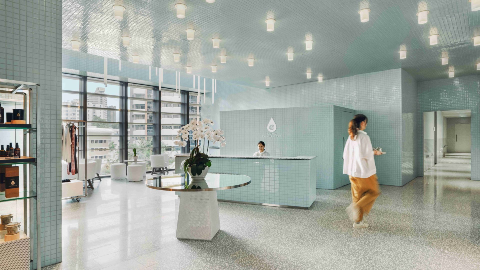 COMO Shambhala Metropolitan, Singapore A light-blue spa reception area with a woman walking toward a doorway. A person sits at the reception desk.