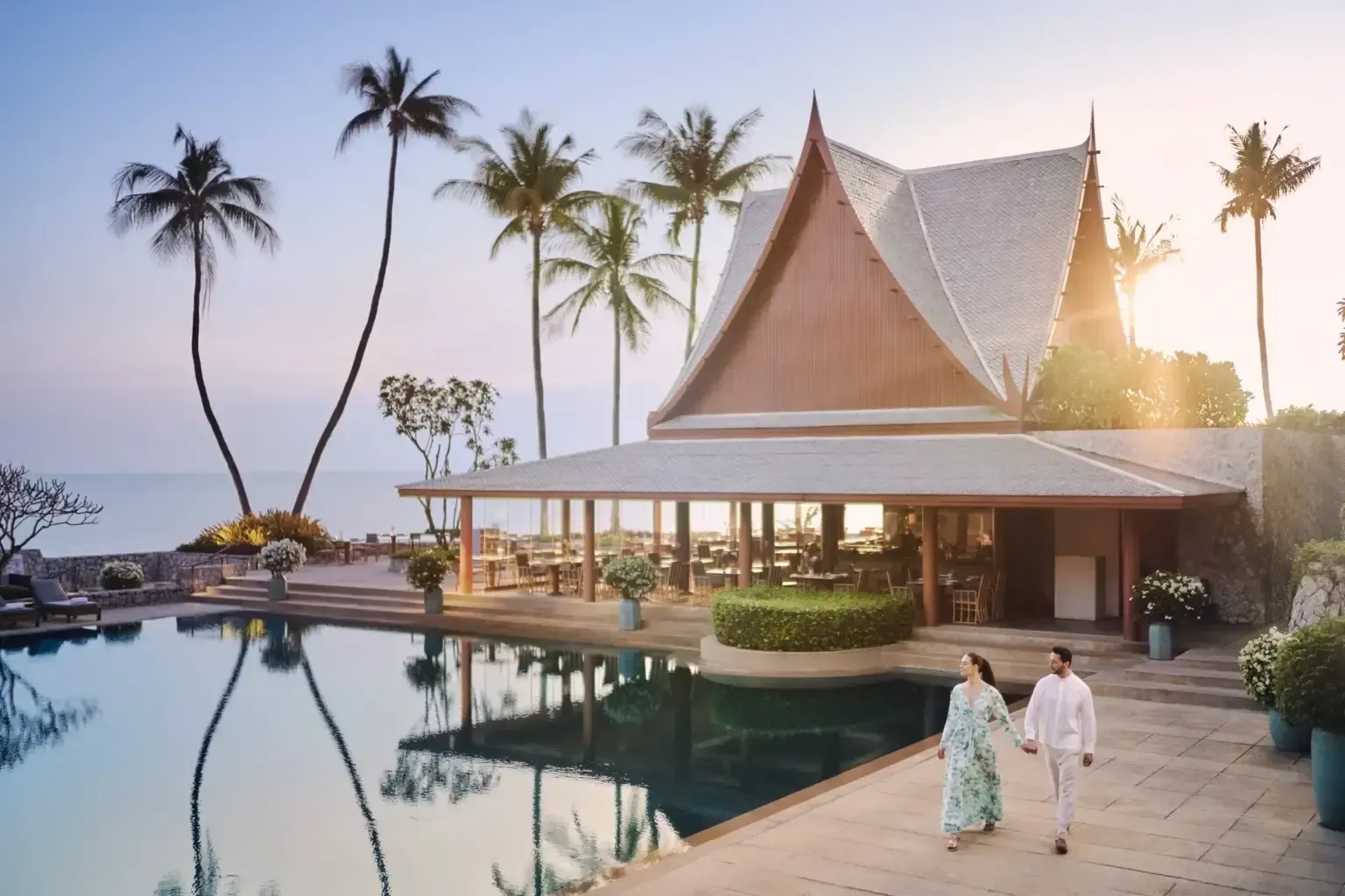 A couple walks hand-in-hand beside a resort pool at dusk, with a traditional Thai-style building and palm trees nearby. Chiva-Som, Hua Hin