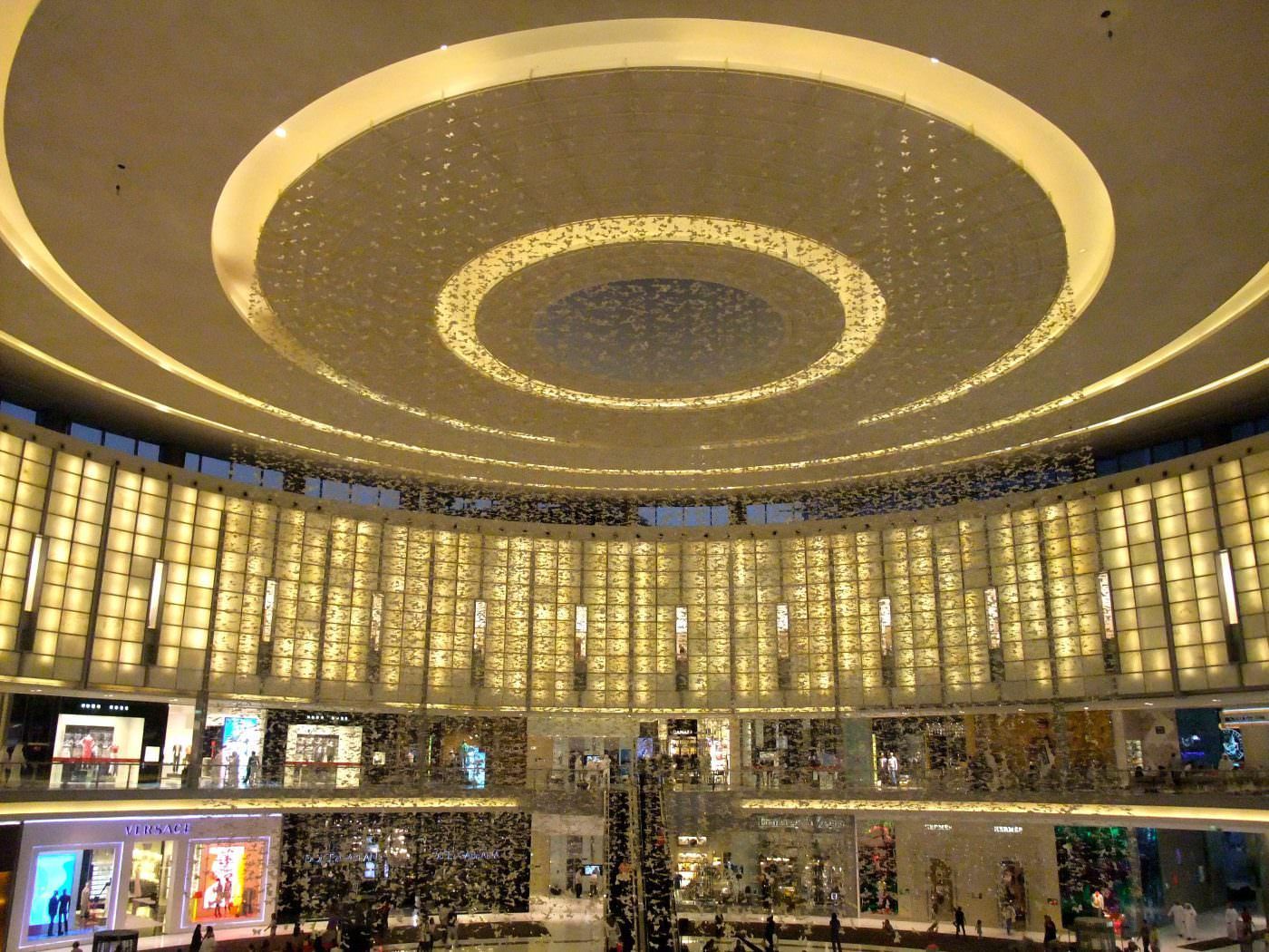 Interior view of a shopping mall atrium with multiple levels, lit by circular ceiling lights.