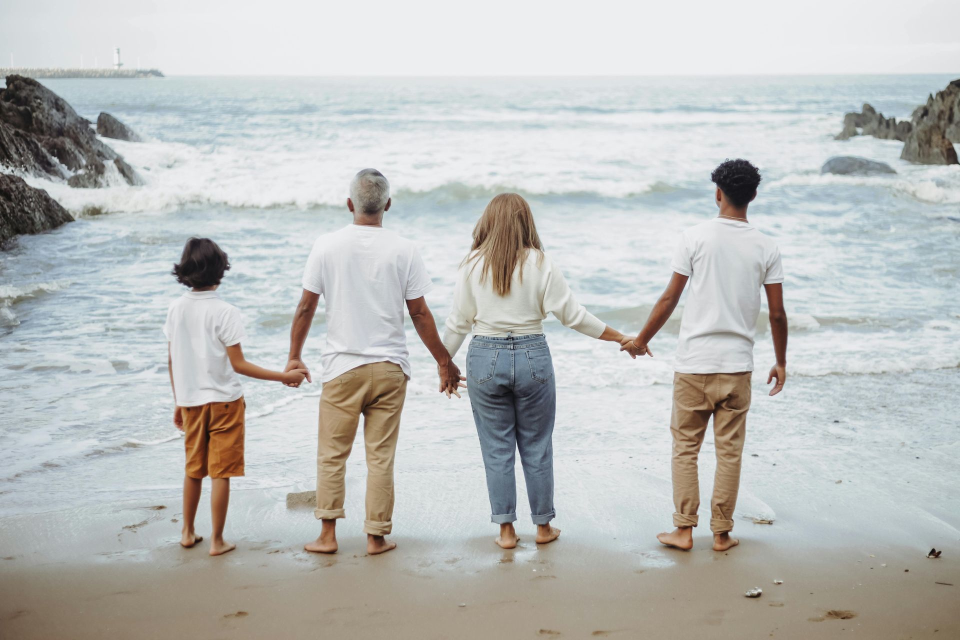 Four people stand in a line on a sandy beach holding hands, looking out at the ocean waves.