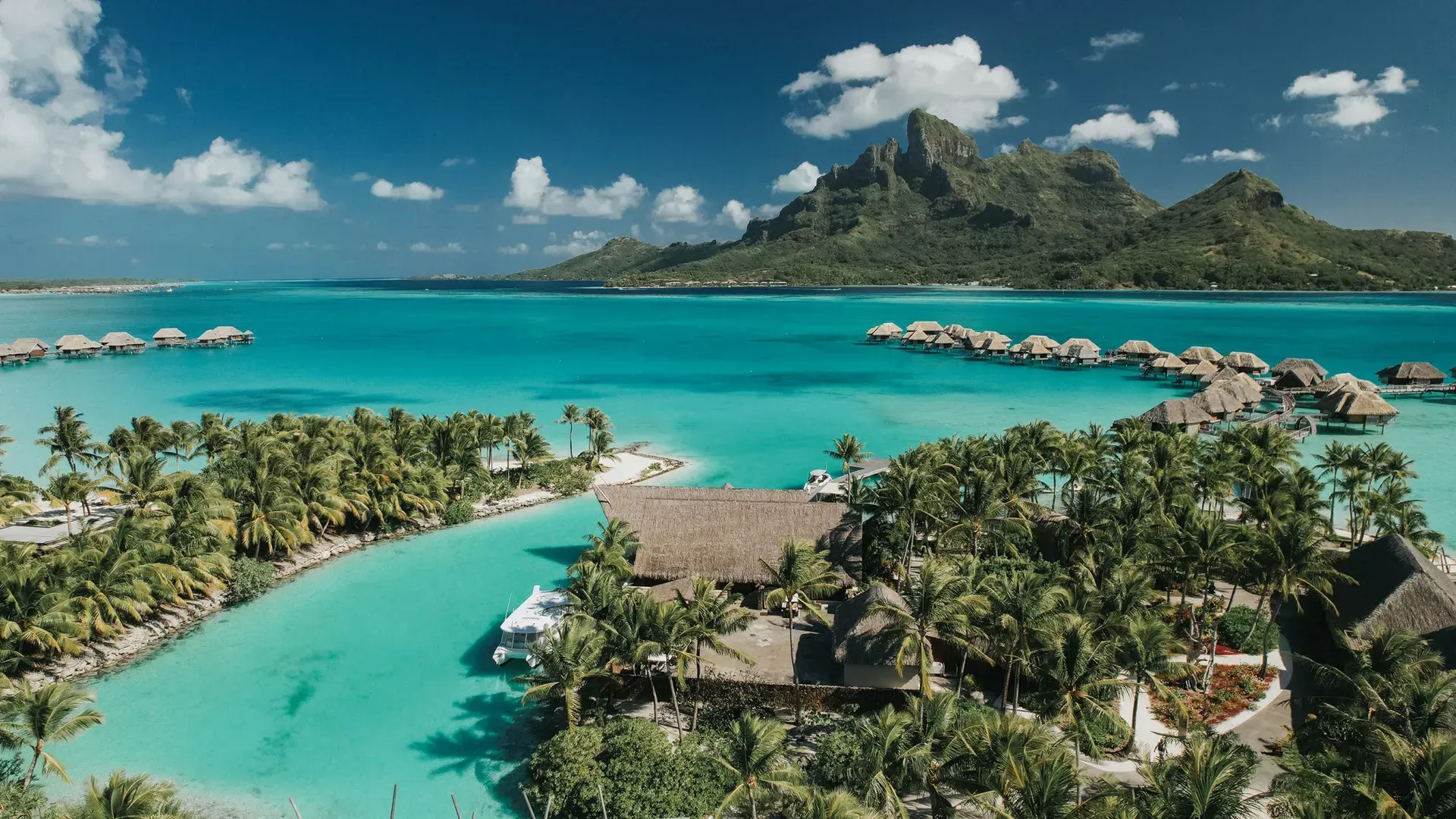 Wooden walkway lit with yellow lights leads to overwater bungalows at dusk. The St. Regis, Bora Bora