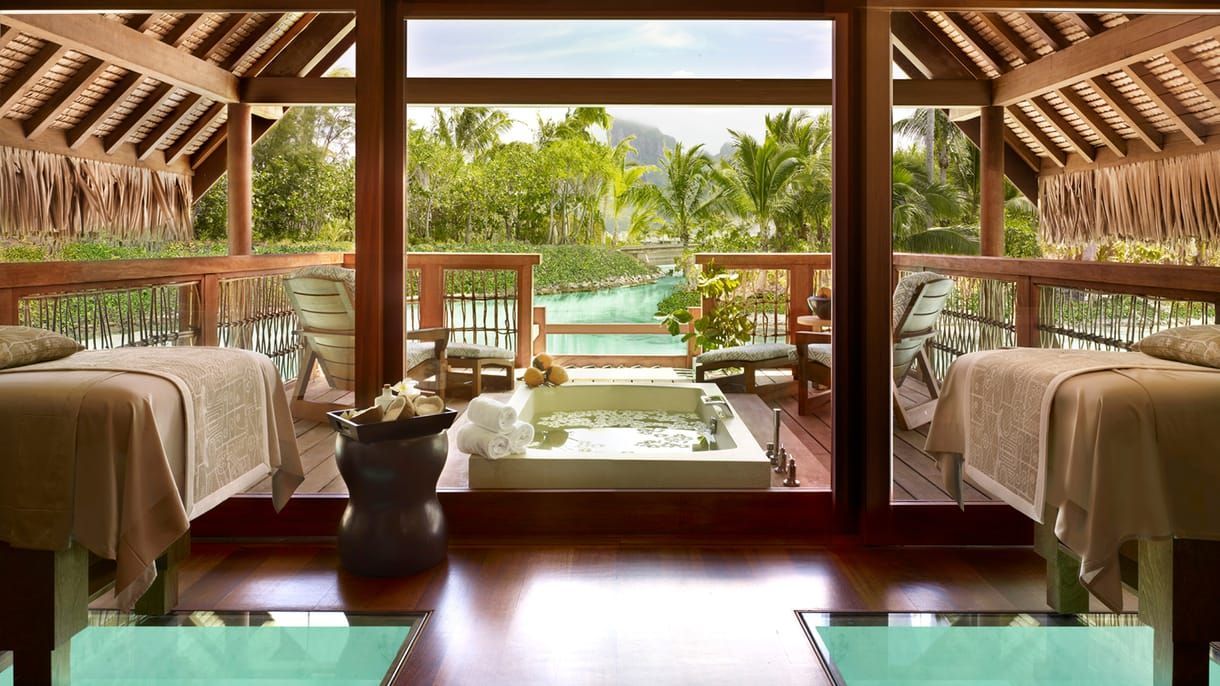 Spa room with two massage tables, a tub, and an ocean view. Wooden structure with a tropical setting. Four Seasons Resort Bora Bora SPA