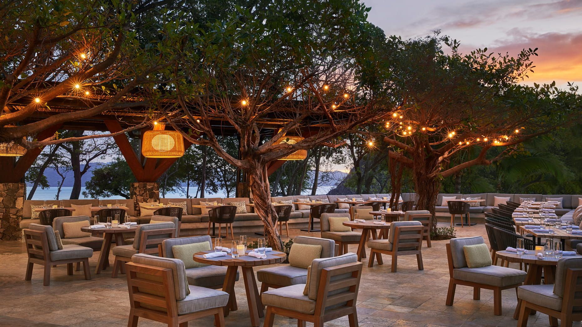 Outdoor restaurant patio with empty wooden tables and chairs under trees lit by warm string lights at dusk. Four Seasons Resort Costa Rica 