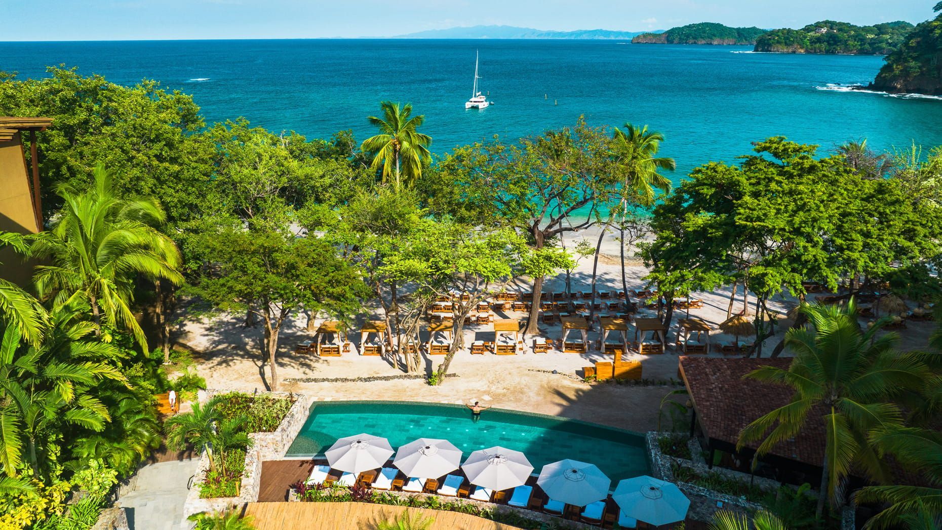 Tropical beach resort with palm trees, blue ocean, sailboat, and white umbrellas by a pool. Four Seasons Resort Costa Rica 
