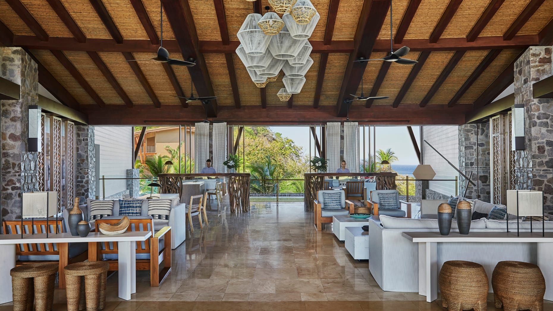 Open-air restaurant with wooden tables, white seating, stone columns, and a hanging chandelier.