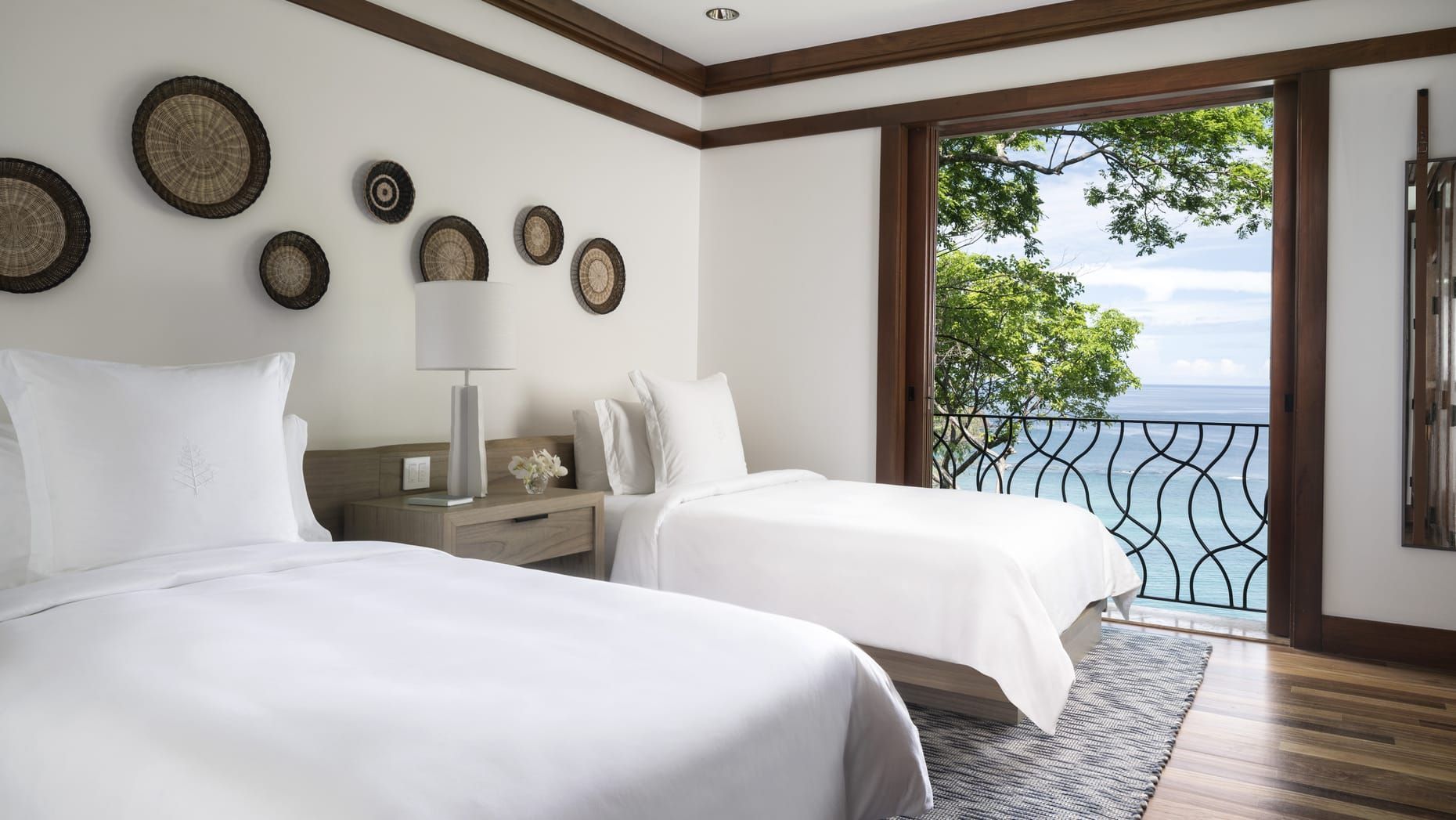 Bright bedroom with two white beds, woven wall decor, and an open balcony overlooking the sea. Four Seasons Resort Costa Rica 