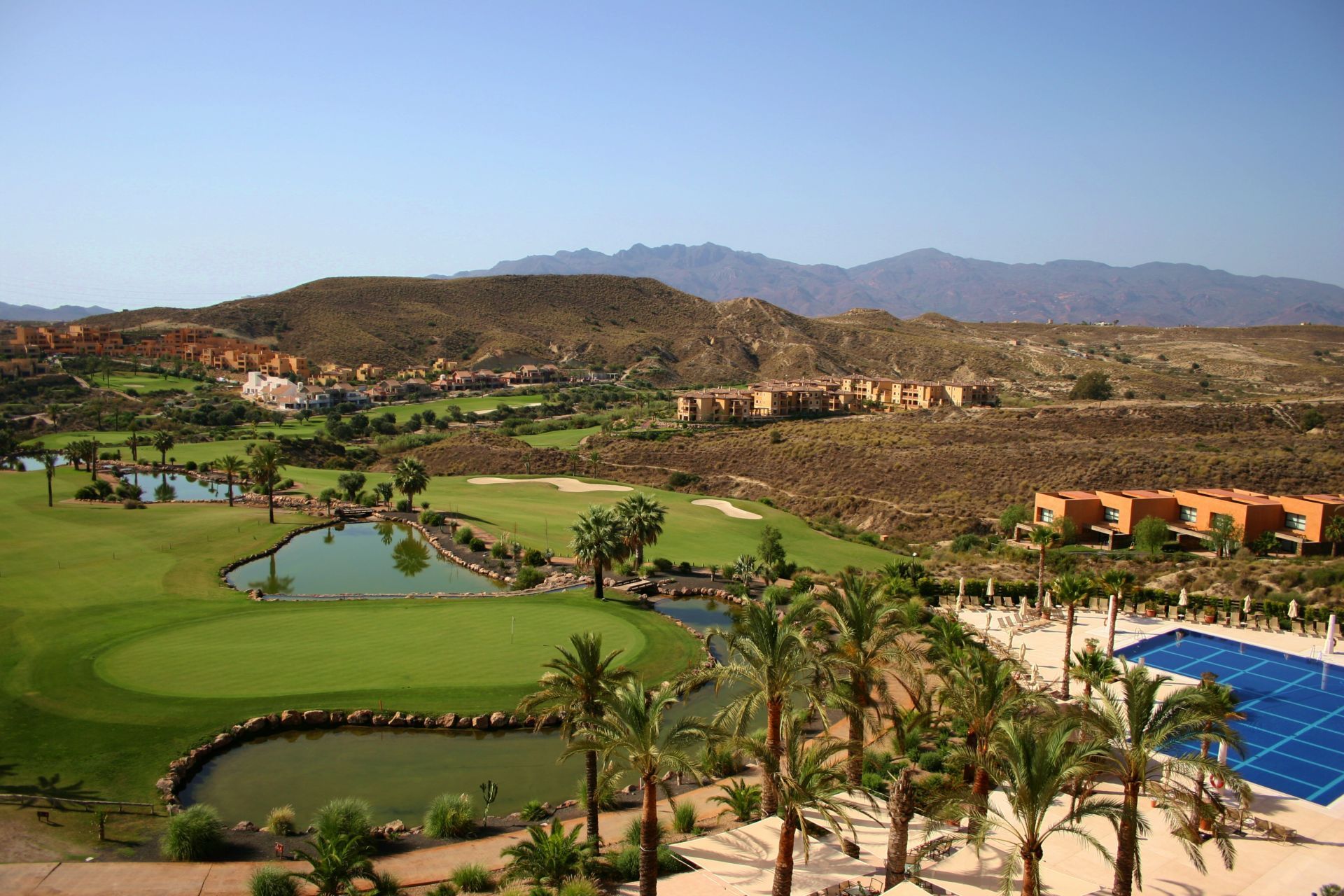 Golf course with green fairways, water features, and palm trees, with distant brown hills and buildings under a blue sky. Spain