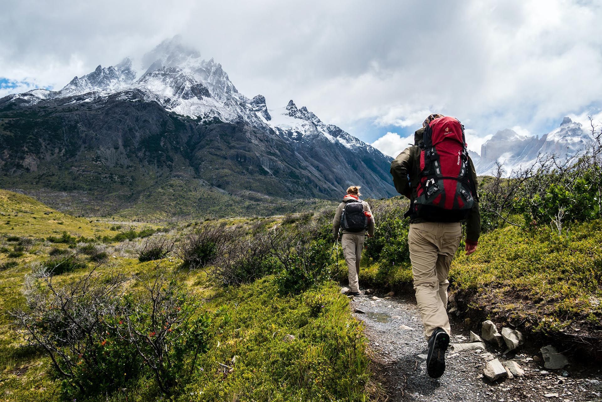 Two hikers trekking a mountain path, snowy peaks in the background, cloudy sky. At Badrutt’s Palace Hotel, 