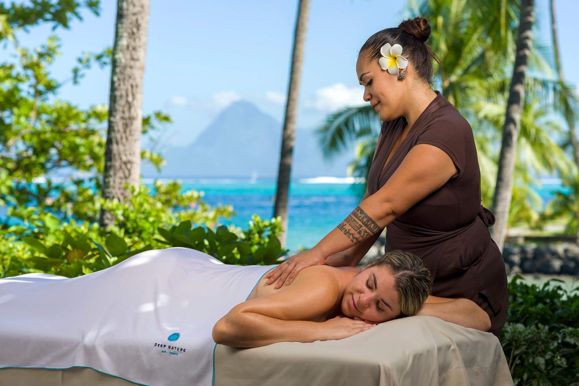 A spa professional performs a massage on a client on a treatment table against a tropical ocean and mountain background. InterContinental, Tahiti spa
