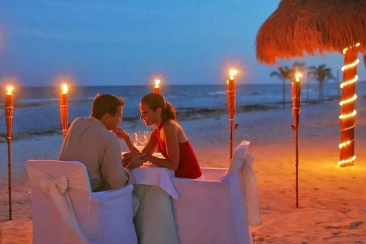 Couple at candlelit beach dinner, looking at each other, torches and palm trees in background. Jimbaran