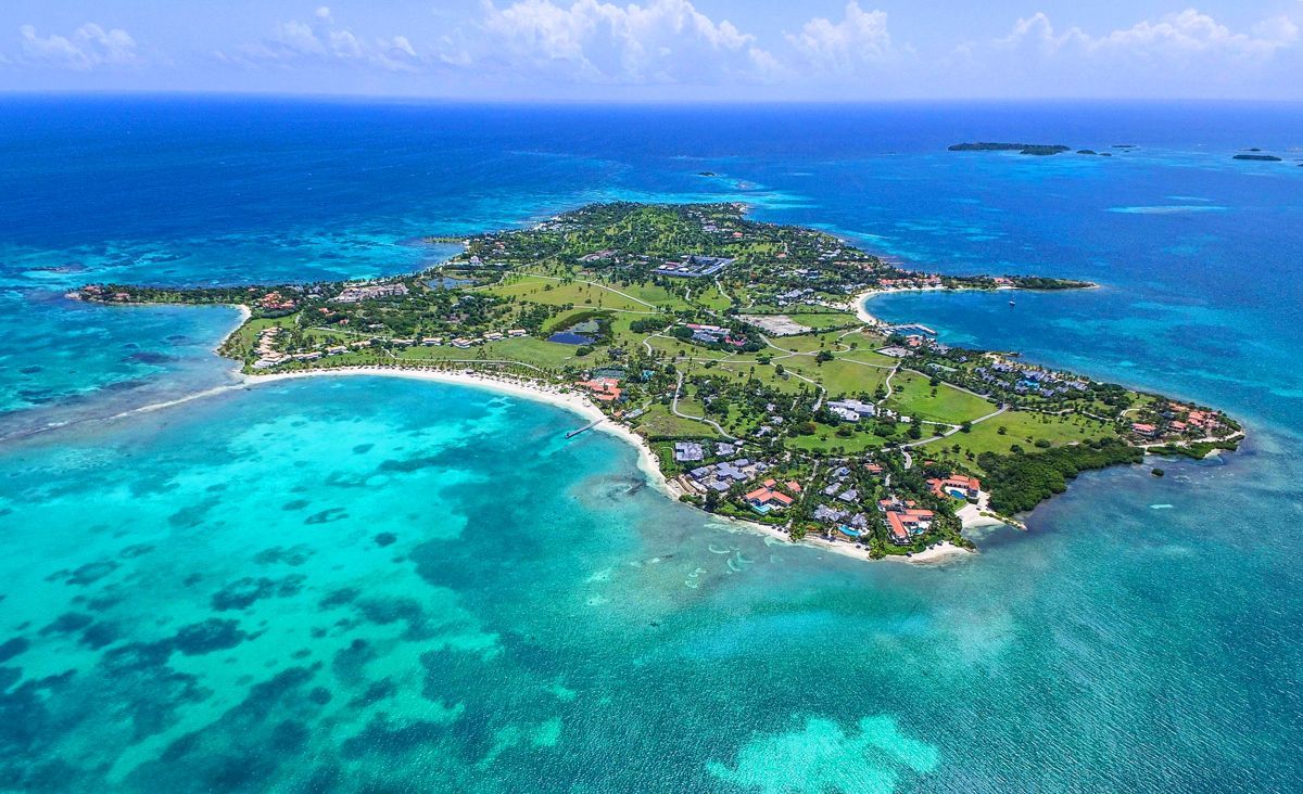 An aerial view of a green tropical island surrounded by turquoise waters with sandy beaches and buildings.