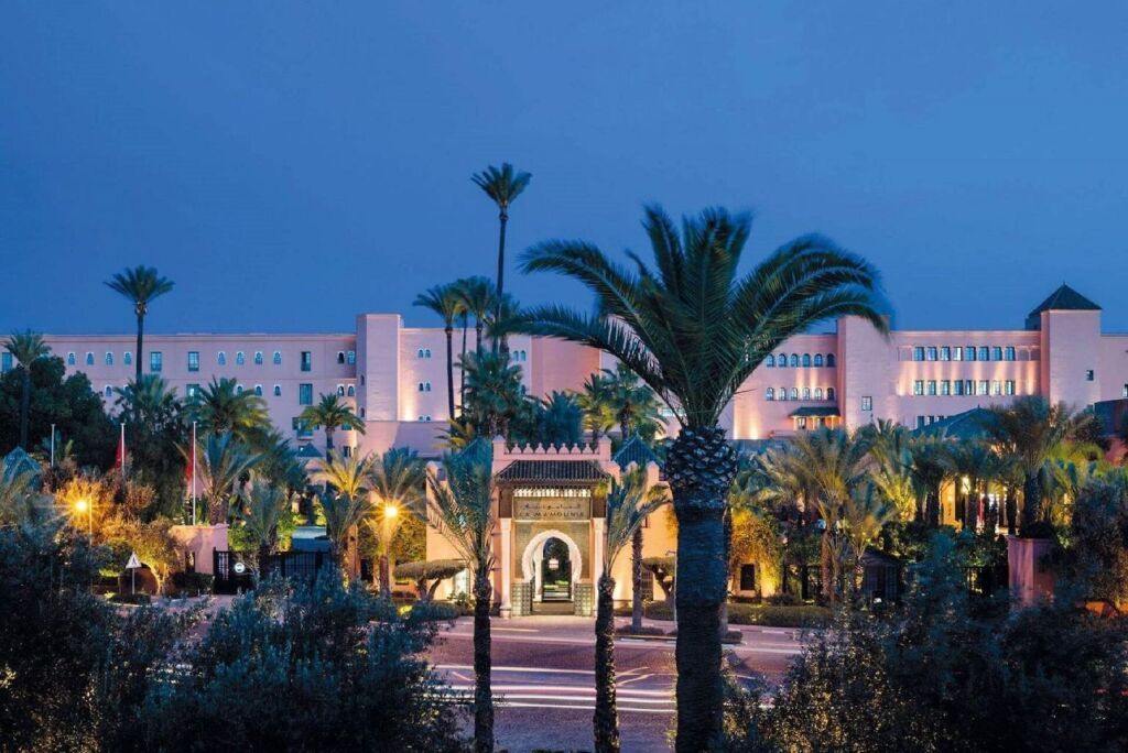 Illuminated resort entrance at dusk with palm trees and a pink-lit building in the background La Mamounia
